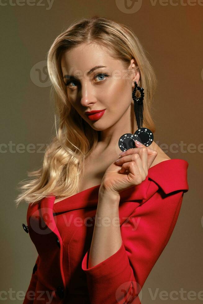 Blonde lady with bright make-up, in red dress and black earrings. She showing two chips, posing on colorful background. Poker, casino. Close-up photo