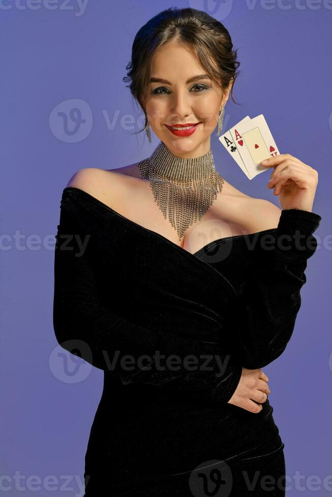 morena dama con desnudo espalda, en negro vestir y brillante joyas. sonriente, demostración dos ases, posando en púrpura antecedentes. póker, casino. de cerca foto