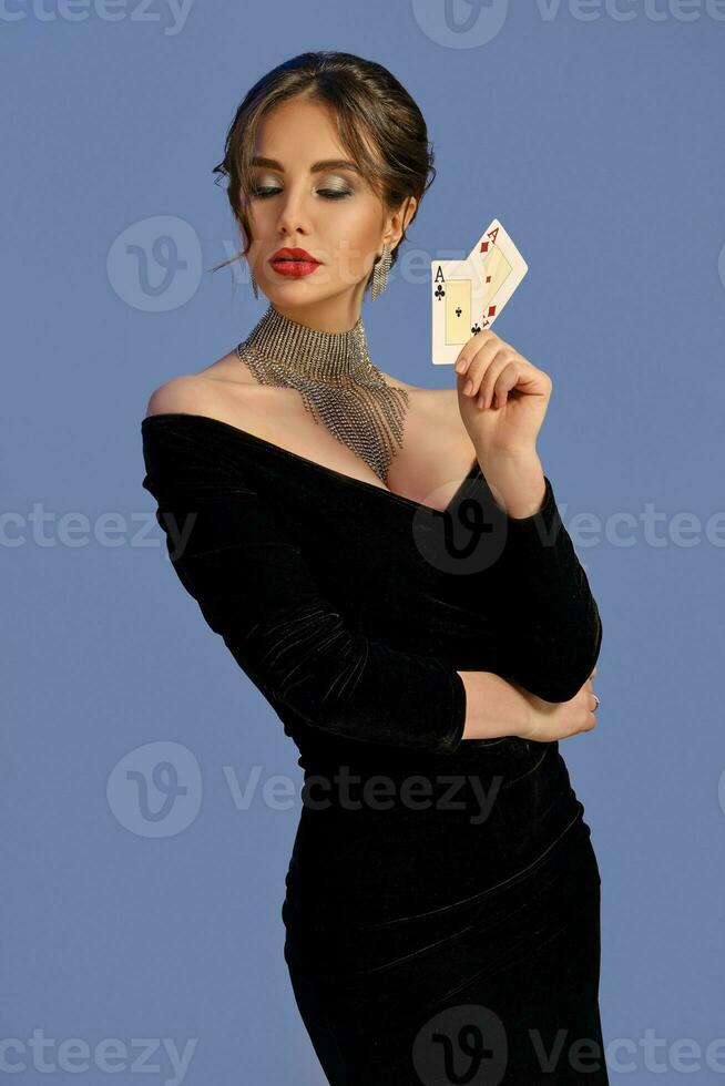 Brunette woman with bare shoulders, in black dress and jewelry. Showing two playing cards, posing on blue background. Poker, casino. Close-up photo