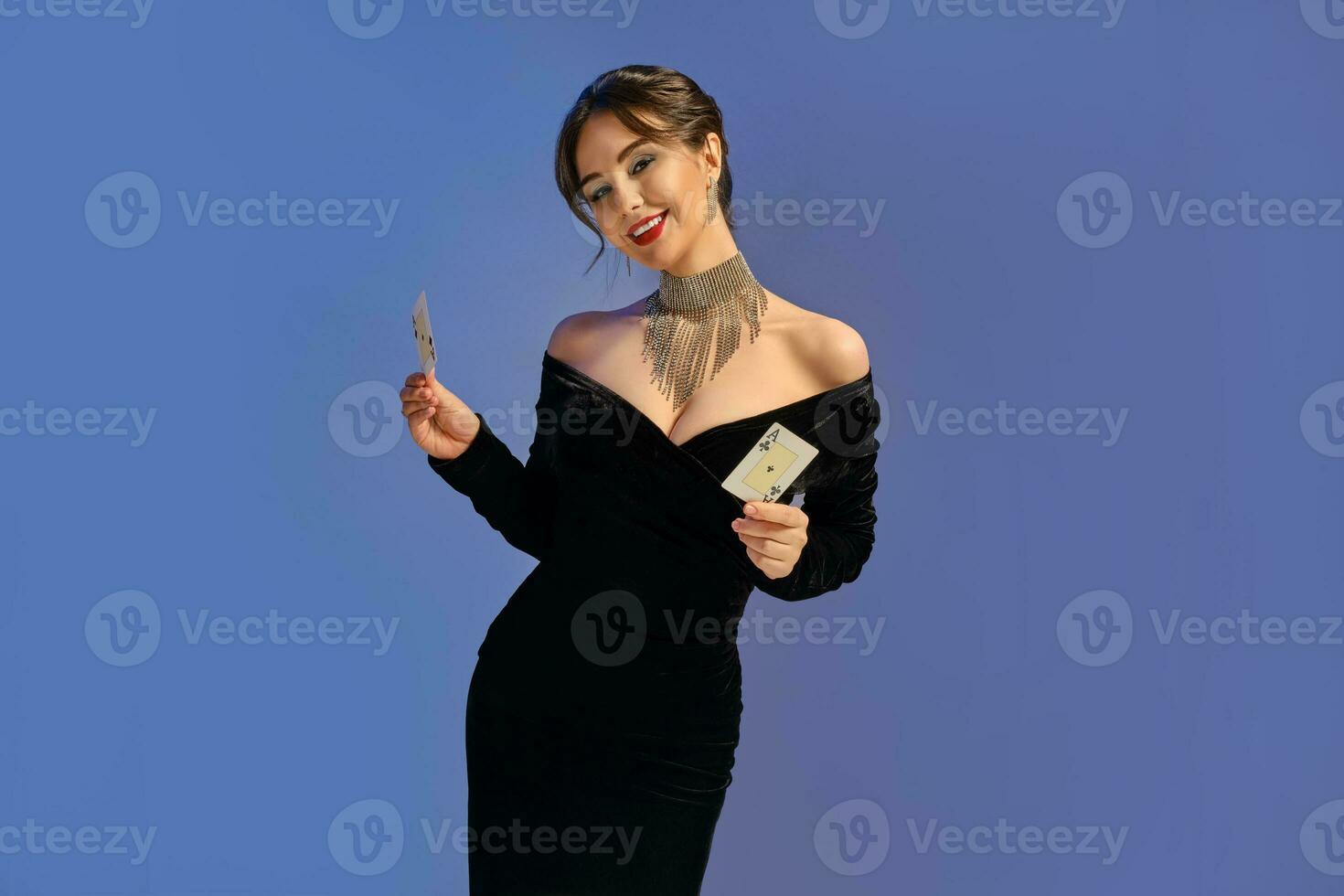 Brunette girl with bare shoulders, in black dress and jewelry. Smiling, showing two playing cards posing on purple background. Poker, casino. Close-up photo