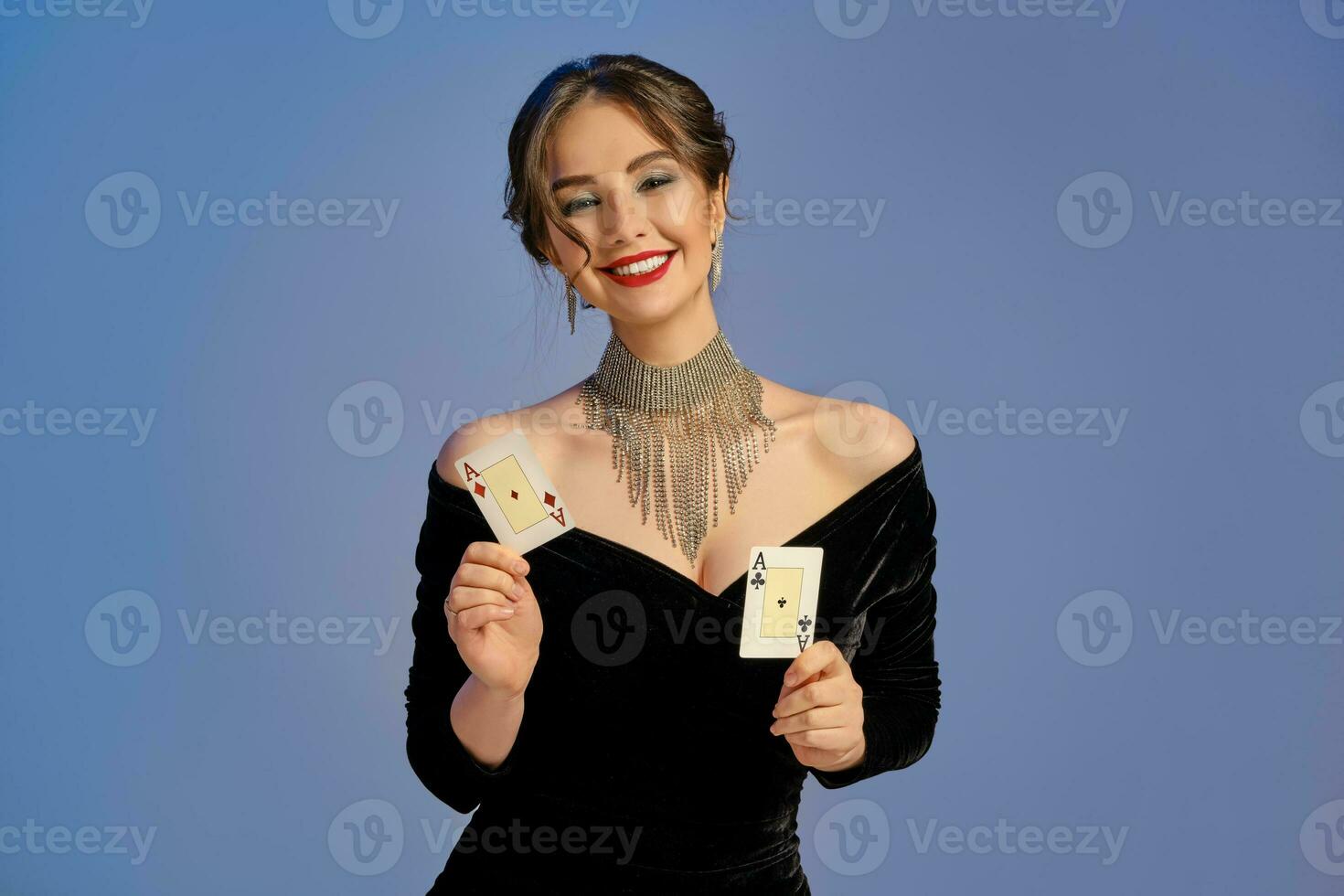 Brunette girl with bare shoulders, in black dress and jewelry. Smiling, showing two playing cards posing on blue background. Poker, casino. Close-up photo