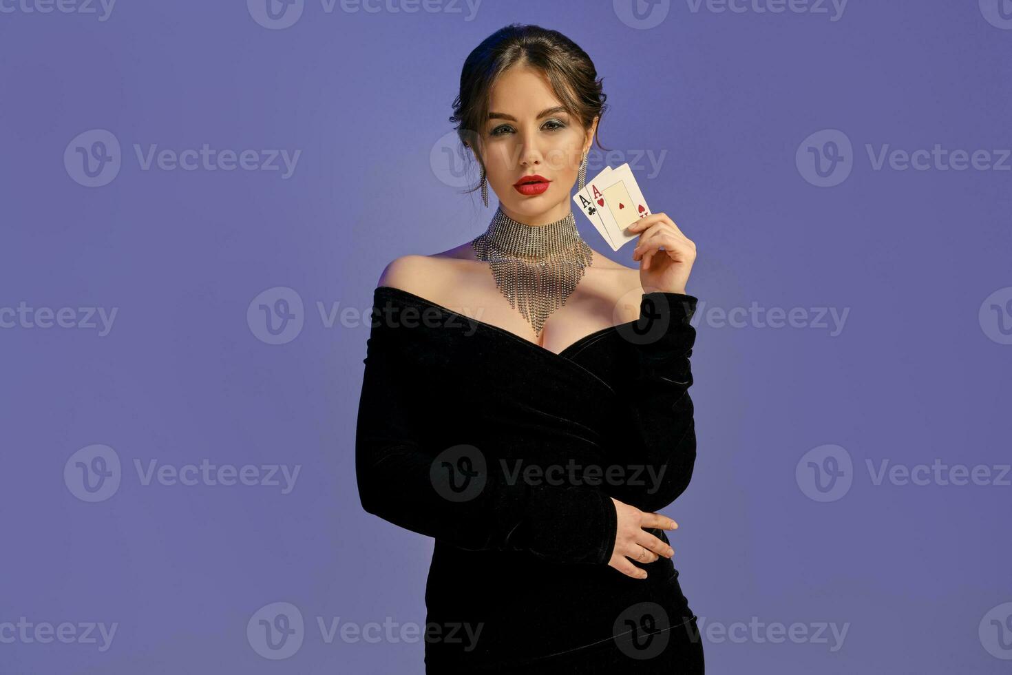 Brunette model with bare shoulders, in black dress and shiny jewelry. Showing two playing cards, posing on purple background. Poker, casino. Close-up photo
