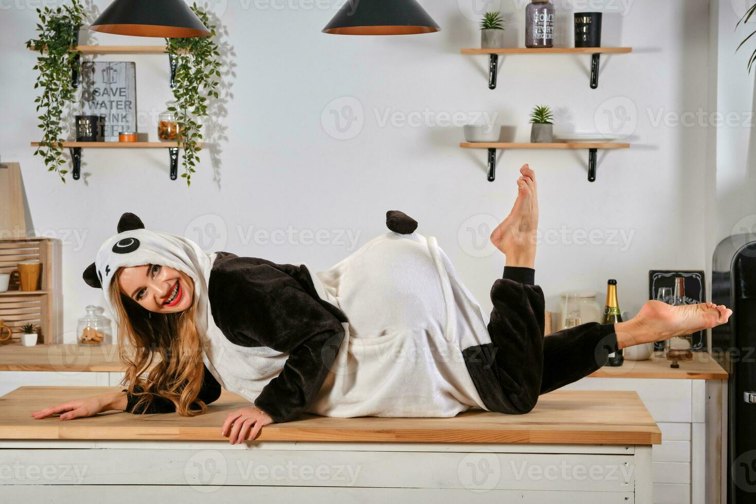 Lady dressed up in plush pajama in form of cartoon character panda is having fun at bachelorette party, posing on kitchen table, smiling. Close-up. photo