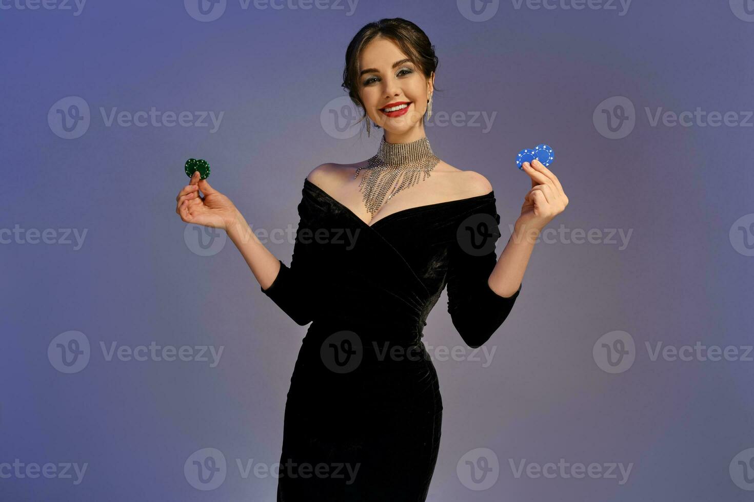 Brunette female in black dress and shiny jewelry. Smiling, showing two blue and green chips, posing on purple background. Poker, casino. Close-up photo