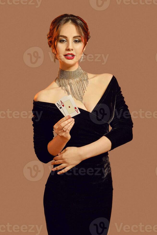 Brunette girl, bare shoulders, in black dress and jewelry. Smiling, showing two playing cards, posing on brown background. Poker, casino. Close-up photo