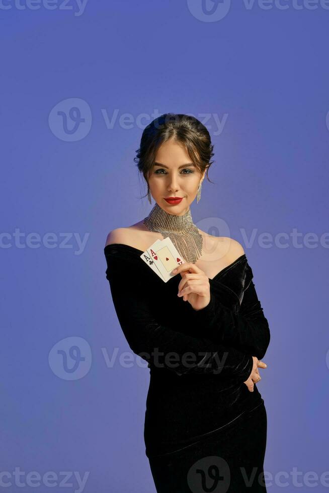 Brunette girl with bare shoulders, in black dress and shiny jewelry. Showing two playing cards, posing on purple background. Poker, casino. Close-up photo