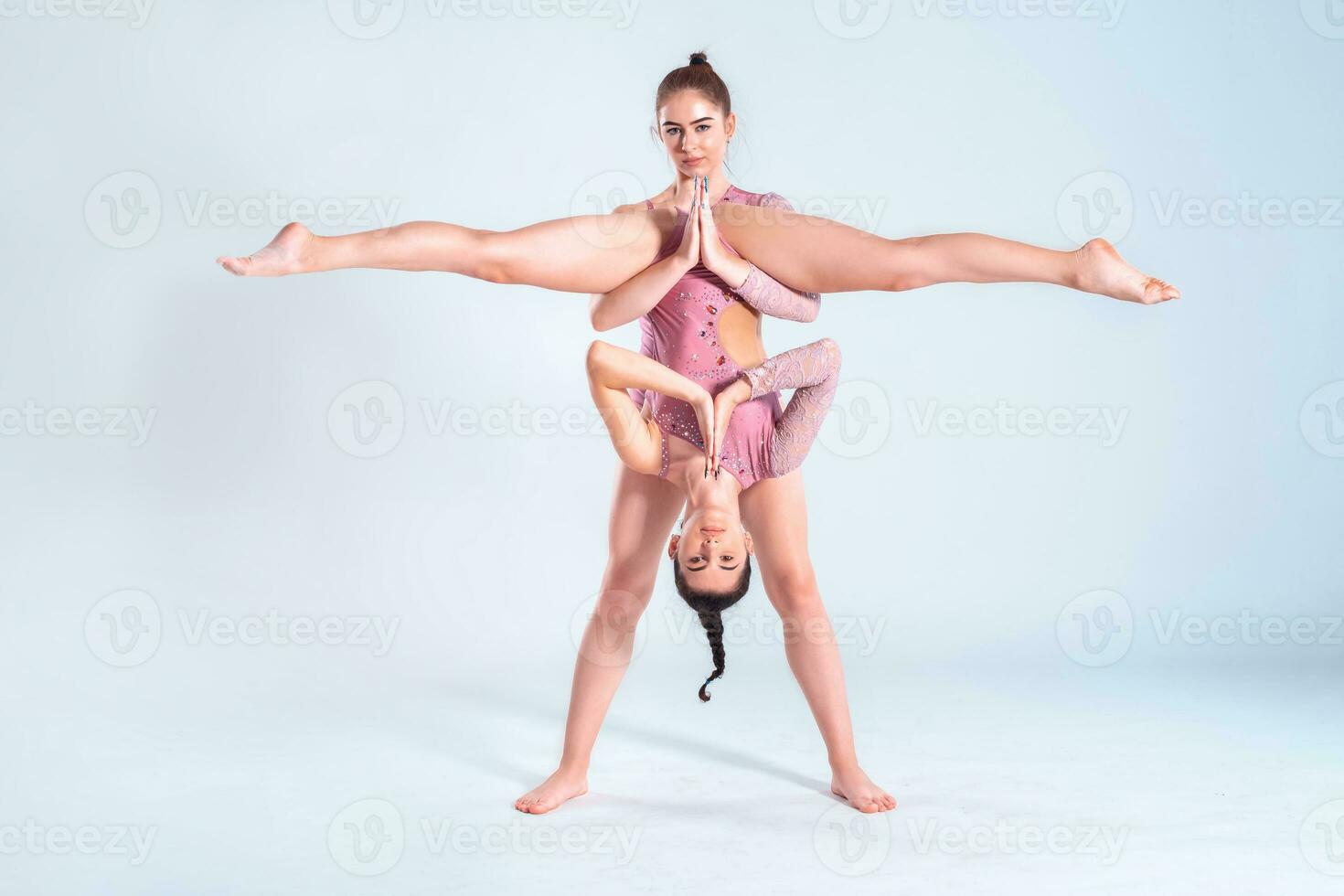 Two flexible girls gymnasts with pigtails, in pink leotards are performing splits using support while posing isolated on white background. Close-up. photo