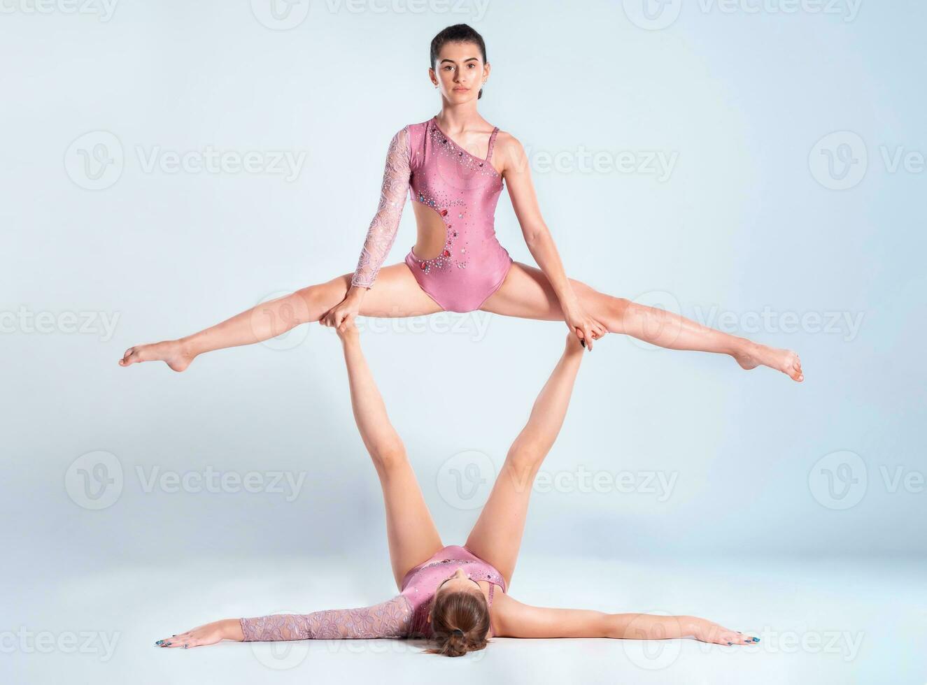 Two flexible girls gymnasts with pigtails, in pink leotards are performing splits using support while posing isolated on white background. Close-up. photo