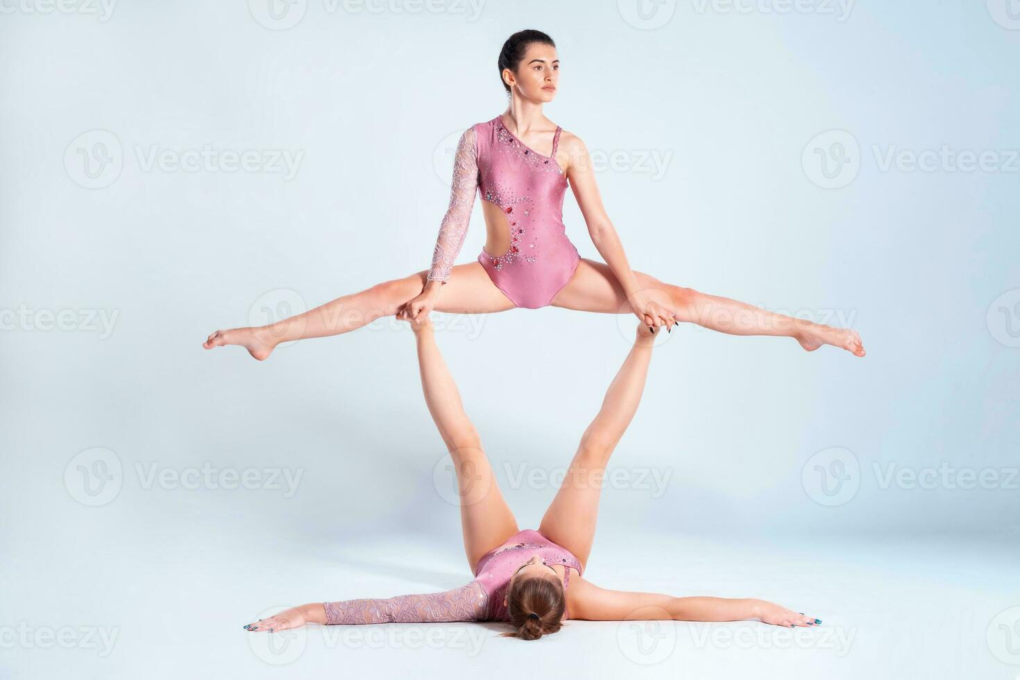 Two flexible girls gymnasts with pigtails, in pink leotards are performing splits using support while posing isolated on white background. Close-up. photo