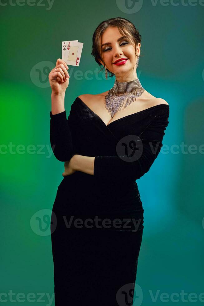 Brunette girl in black dress, necklace and earrings. Smiling, showing two playing cards, posing on colorful background. Poker, casino. Close-up photo