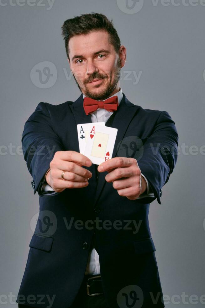 Man in black classic suit and red bow-tie showing two playing cards while posing against gray studio background. Gambling, poker, casino. Close-up. photo