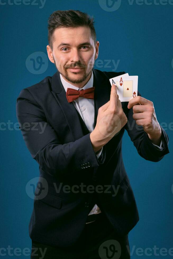 Man in black classic suit and red bow-tie showing two playing cards while posing against blue studio background. Gambling, poker, casino. Close-up. photo