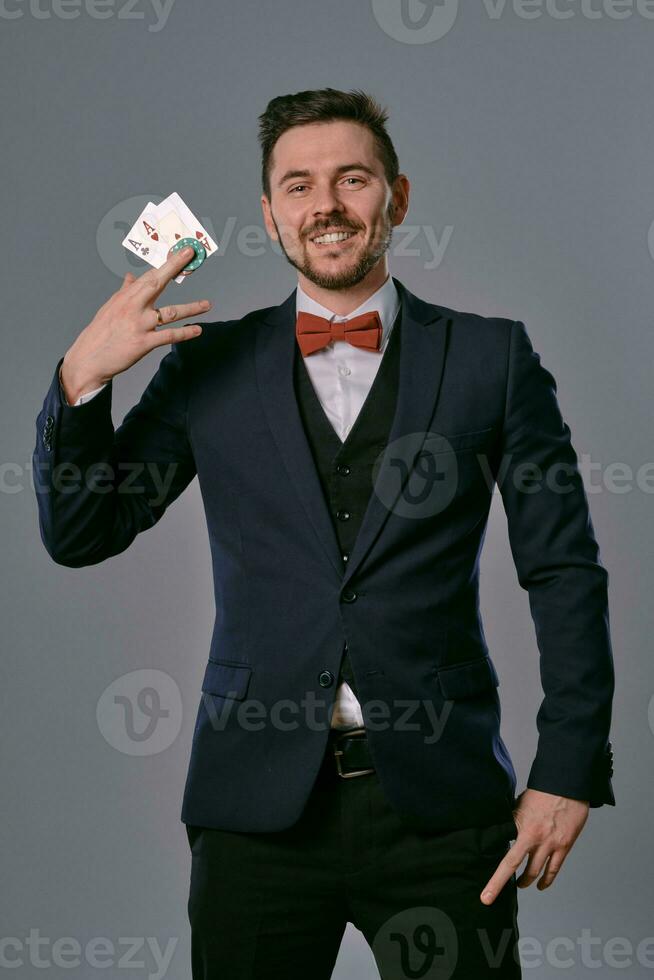 Man in black classic suit and red bow-tie showing two playing cards and chips, posing on gray studio background. Gambling, poker, casino. Close-up. photo