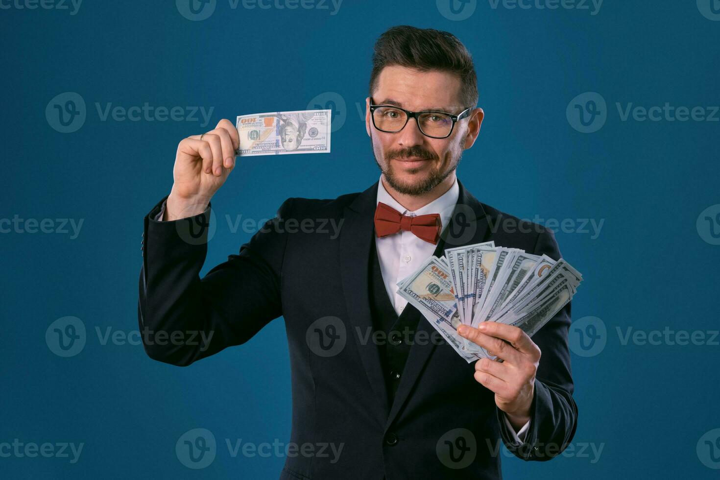 hombre en negro clásico traje, rojo corbata de moño, glases es participación algunos dólar facturas, posando en gris estudio antecedentes. juego, póker, casino. de cerca. foto