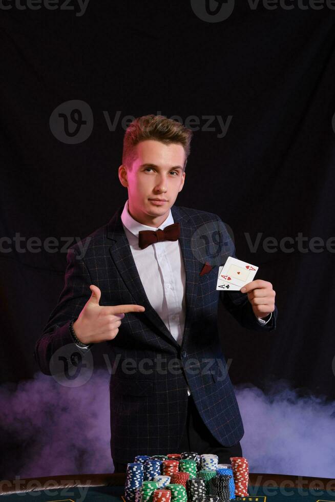 elegante hombre es jugando póker a casino. él es posando a el mesa con pilas de de colores papas fritas en él, en contra un negro, fumar antecedentes. foto