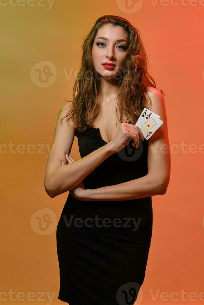 Brunette female with earring in nose, in black dress. Showing two playing cards, posing on colorful background. Poker, casino. Close-up photo