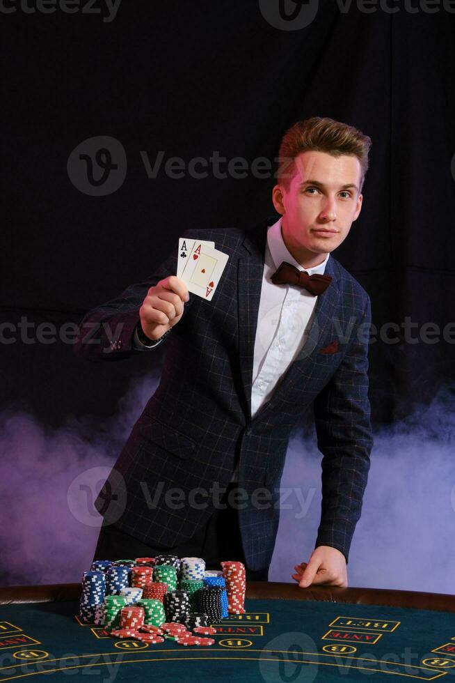 Elegant man playing poker at casino. He is showing two aces while posing at the table with stacks of chips on it, against black background. photo