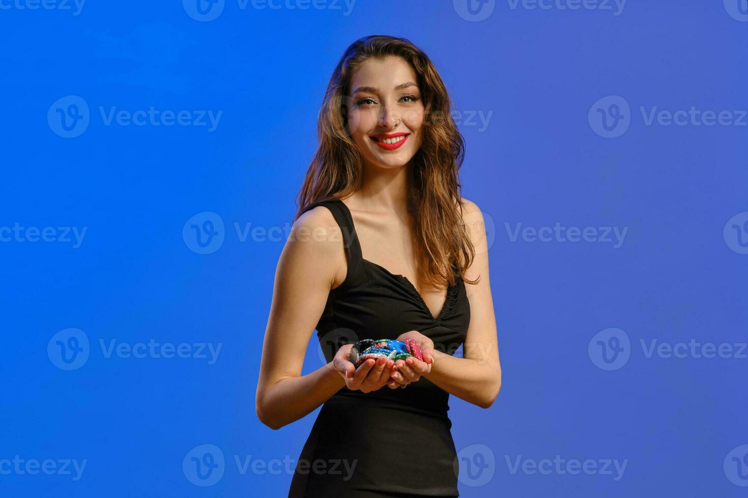 Brunette model in black dress. She is smiling, showing handful of colorful chips, posing on blue background. Poker, casino. Close-up photo