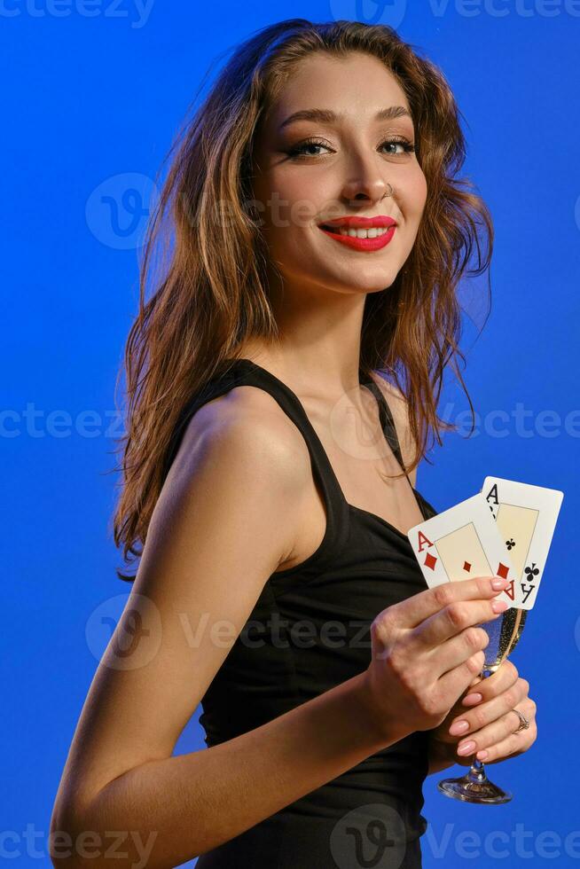 Brunette female in black dress. Holding glass of champagne, two aces, smiling, posing sideways on blue background. Poker, casino. Close-up photo