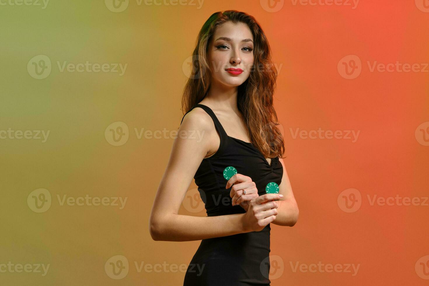 Brunette model with earring in nose, in black dress. Holding two green chips, posing sideways on colorful background. Poker, casino. Close-up photo