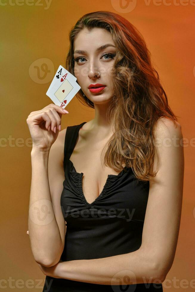 Brunette model with earring in nose, in black dress. She is showing two aces, posing on colorful studio background. Gambling, poker, casino. Close-up photo