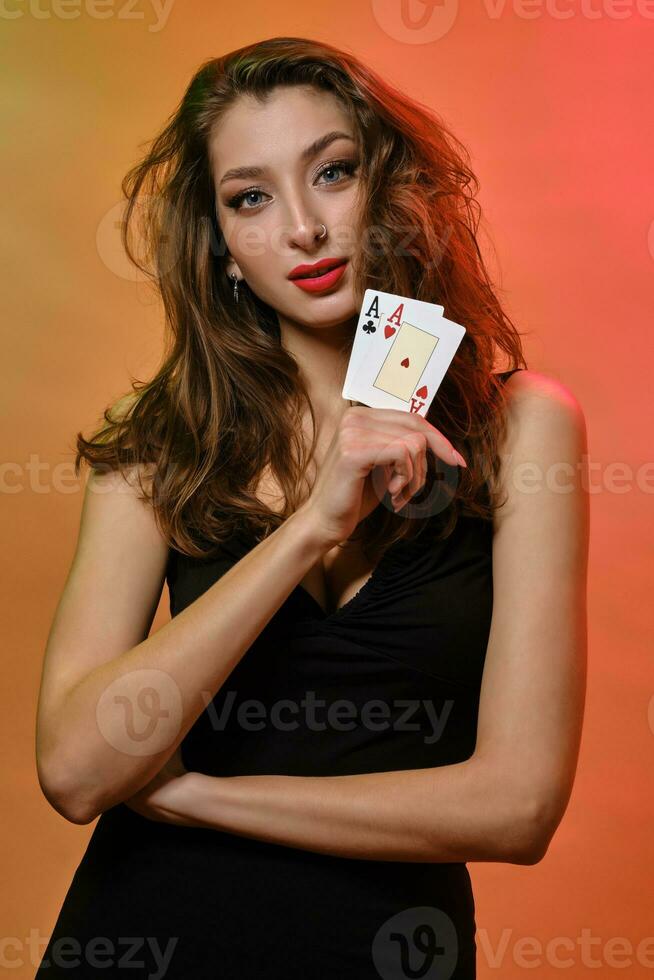 Brunette girl with earring in nose, in black dress. Showing two aces, posing on colorful background. Gambling, poker, casino. Close-up photo