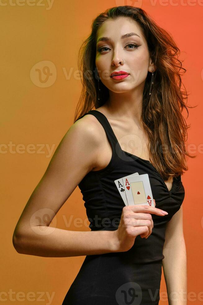 Brunette lady with earring in nose, in black dress. She showing two playing cards, posing sideways on colorful background. Poker, casino. Close-up photo