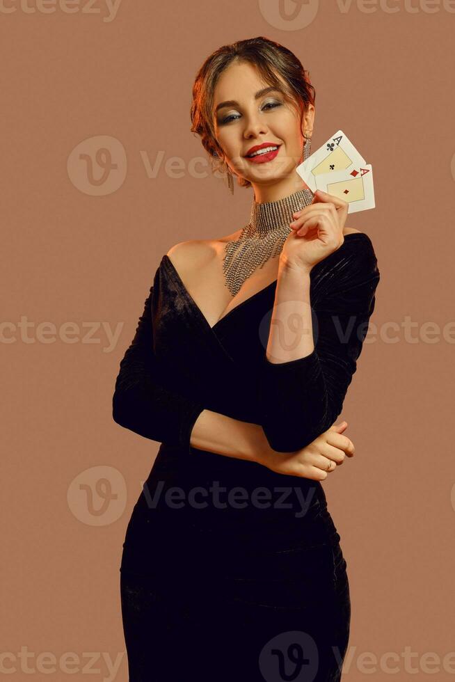 Brunette woman, bare shoulders, in black dress and jewelry. Smiling, showing two playing cards, posing on brown background. Poker, casino. Close-up photo