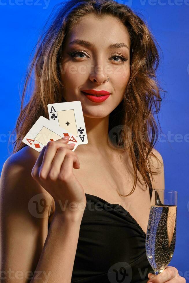 Brunette model with earring in nose, in black dress. Holding glass of champagne, two aces, smiling, posing on blue background. Poker, casino. Close-up photo