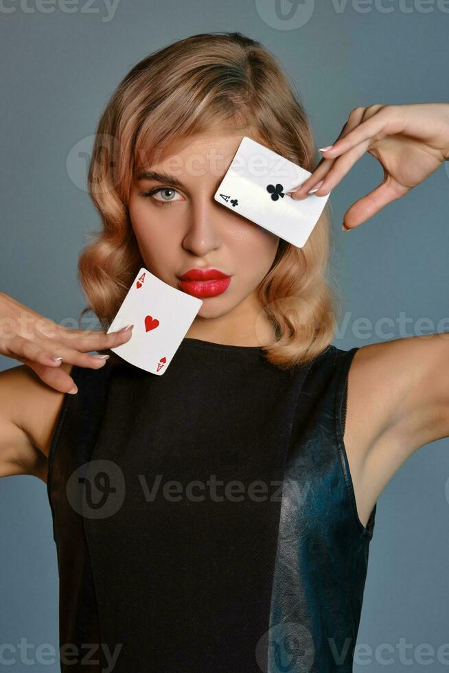 Blonde girl in black leather dress showing two playing cards, posing against gray background. Gambling entertainment, poker, casino. Close-up. photo