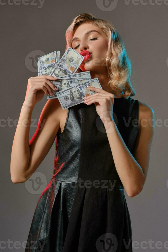 rubia niña en negro elegante vestir participación y besos algunos dinero, posando en contra gris antecedentes. juego entretenimiento, póker, casino. de cerca. foto