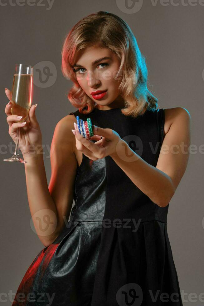 Blonde girl in black dress holding glass of champagne and chips, posing against gray background. Gambling, poker, casino. Close-up. photo