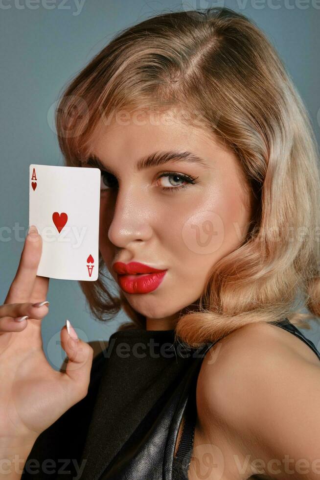 Blonde girl in black leather dress showing ace of hearts, posing against gray background. Gambling entertainment, poker, casino. Close-up. photo