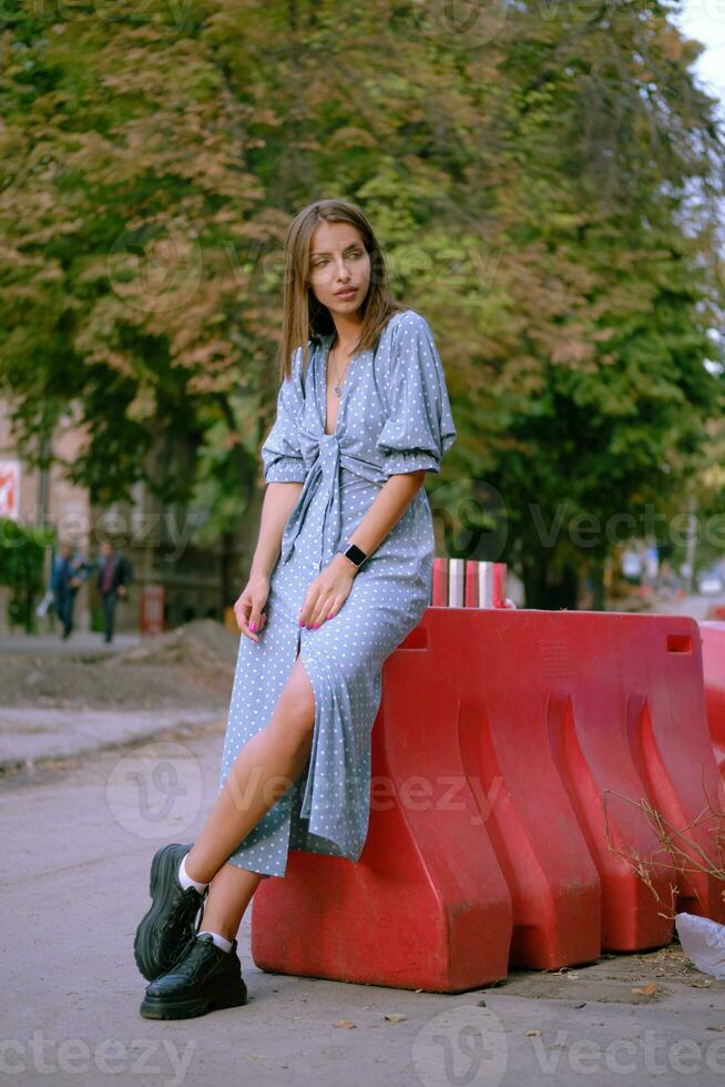 rubia niña en largo azul vestir y un pequeño negro bolso en su hombro es posando cerca un rojo Barandilla, caminando solo en el ciudad. lleno longitud disparo. foto