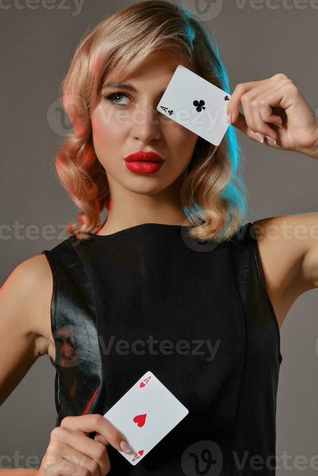 Blonde girl in black leather dress showing two playing cards, posing against gray background. Gambling entertainment, poker, casino. Close-up. photo