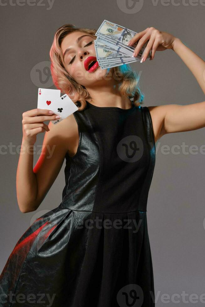 Blonde girl in black stylish dress holding some money and cards, posing against gray background. Gambling entertainment, poker, casino. Close-up. photo