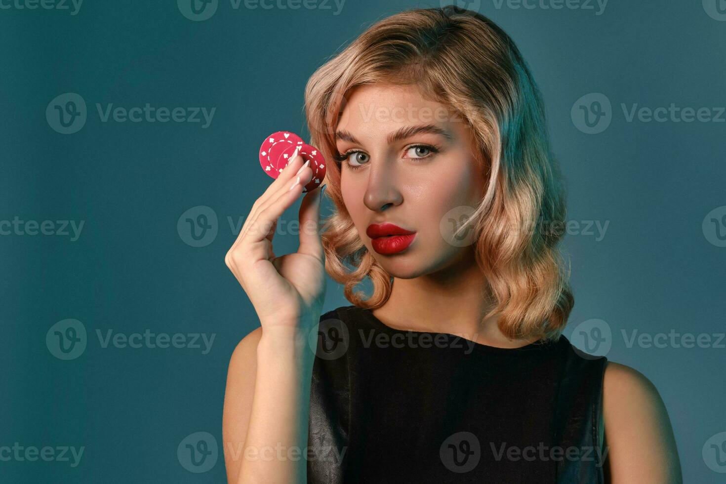 Blonde girl in black leather dress showing two red chips, posing against blue background. Gambling entertainment, poker, casino. Close-up. photo