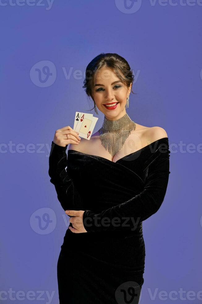 Brunette woman with bare shoulders, in black dress and shiny jewelry. Smiling, showing two aces, posing on purple background. Poker, casino. Close-up photo