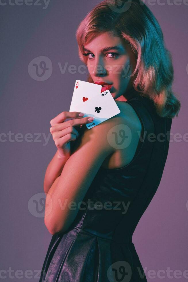 Blonde girl in black stylish dress showing two playing cards, posing against colorful background. Gambling entertainment, poker, casino. Close-up. photo