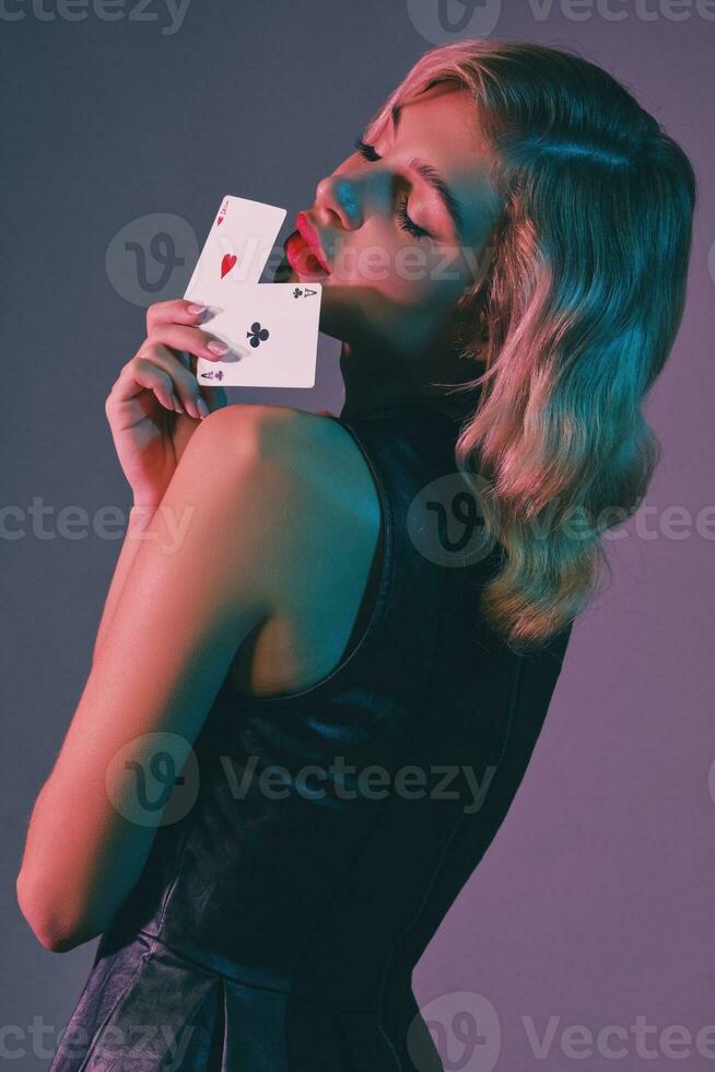 Blonde girl in black stylish dress showing two playing cards, posing against colorful background. Gambling entertainment, poker, casino. Close-up. photo