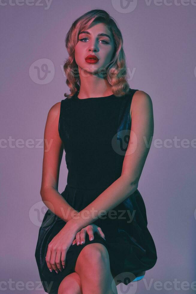 Blonde girl with bright make-up, in black stylish dress is posing sitting on chair against colorful studio background. Fashion and beauty. Close-up. photo