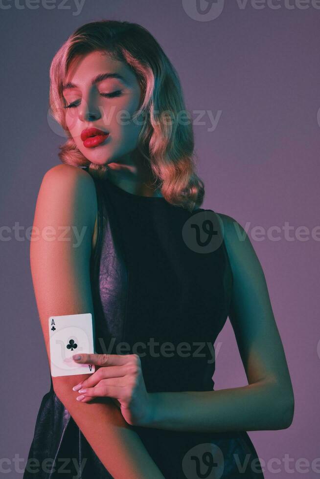 Blonde girl in black stylish dress showing ace of clubs, posing against colorful background. Gambling entertainment, poker, casino. Close-up. photo