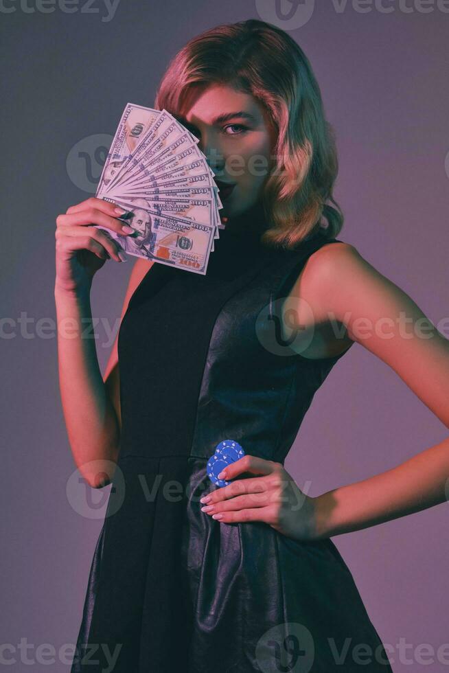 Blonde girl in black stylish dress holding some money and chips, posing against colorful background. Gambling entertainment, poker, casino. Close-up. photo