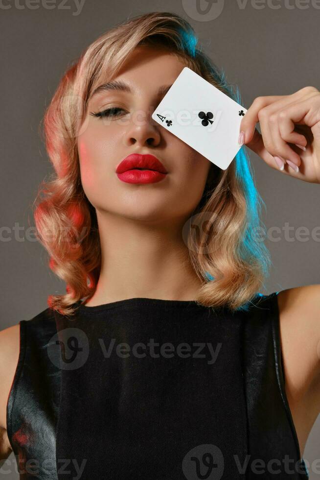 Blonde girl in black leather dress showing ace of clubs, posing against gray background. Gambling entertainment, poker, casino. Close-up. photo