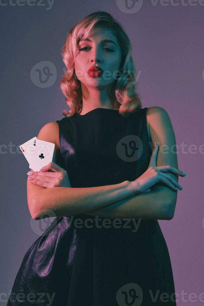 Blonde girl in black stylish dress showing two playing cards, posing against colorful background. Gambling entertainment, poker, casino. Close-up. photo