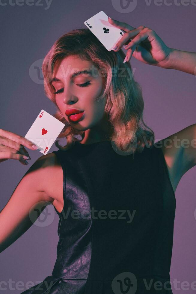 Blonde girl in black stylish dress showing two playing cards, posing against colorful background. Gambling entertainment, poker, casino. Close-up. photo