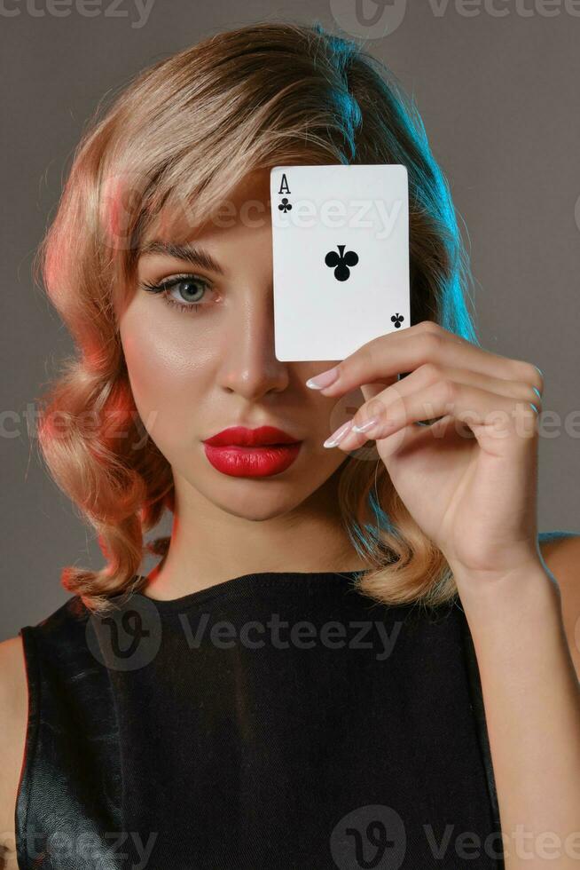 Blonde girl in black leather dress showing ace of clubs, posing against gray background. Gambling entertainment, poker, casino. Close-up. photo