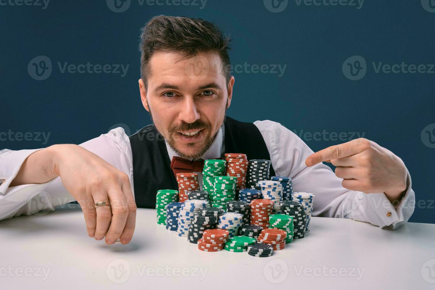 Man in black vest and shirt sitting at white table with stacks of chips on it, posing on blue studio background. Gambling, poker, casino. Close-up. photo