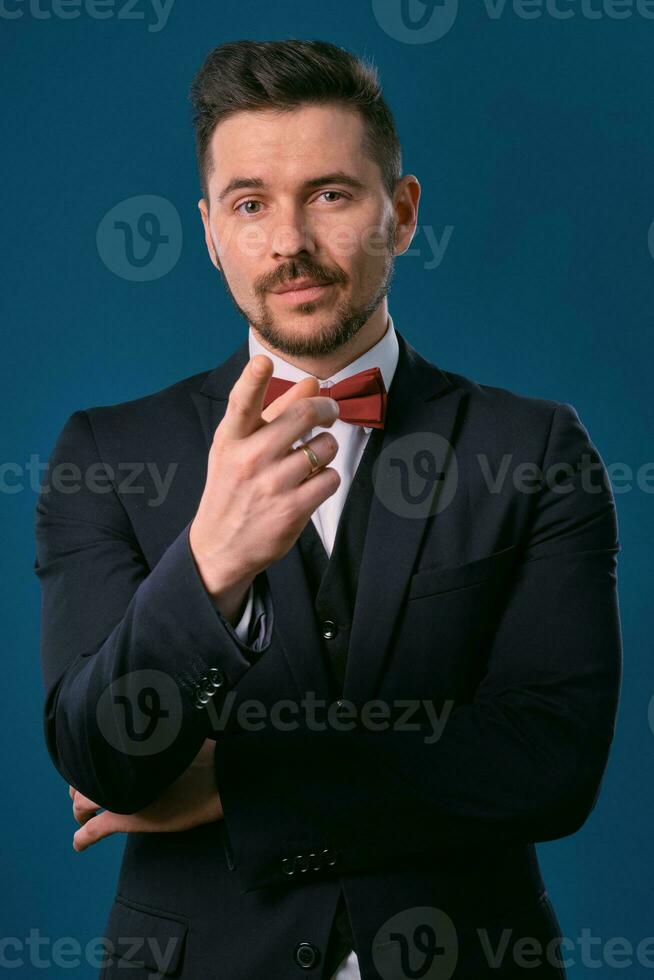 Young business man in classic black suit, white shirt and red bow-tie. Posing against a blue studio background. Mock up, copy space. Close-up. photo