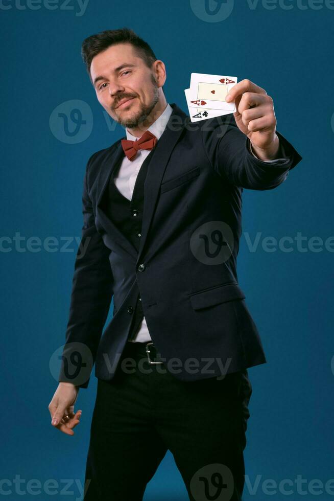 Man in black classic suit and red bow-tie showing two playing cards while posing against blue studio background. Gambling, poker, casino. Close-up. photo